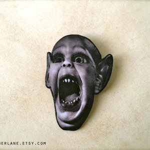 May include: A black and white pin featuring a close-up of a screaming, grotesque, child-like face with large ears.