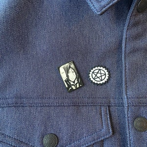 May include: A black and white enamel pin featuring a character from the anime series Black Butler. The character is a man with black hair and a black suit. The pin is attached to a blue denim jacket. There is also a black and white pentagram pin attached to the jacket.