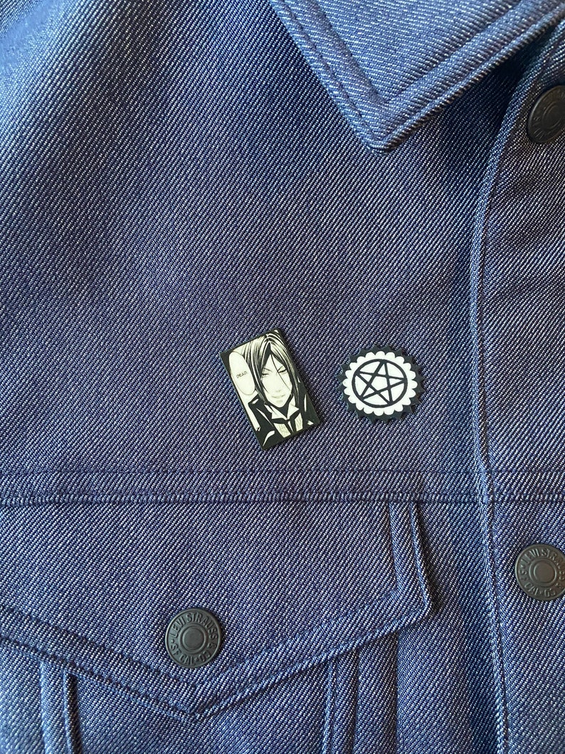 May include: A black and white enamel pin featuring a character from the anime series Black Butler. The character is a man with black hair and a black suit. The pin is attached to a blue denim jacket. There is also a black and white pentagram pin attached to the jacket.