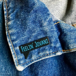 Puede incluir: Un pin de esmalte negro con forma de bocadillo de di&aacute;logo y el texto "Feelin' Jovani" en letras azules. El pin est&aacute; sujeto a una chaqueta vaquera azul.