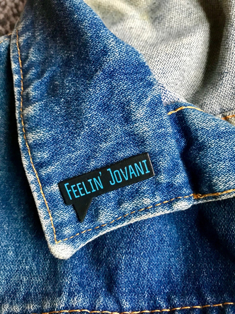 Puede incluir: Un pin de esmalte negro con forma de bocadillo de di&aacute;logo y el texto "Feelin' Jovani" en letras azules. El pin est&aacute; sujeto a una chaqueta vaquera azul.