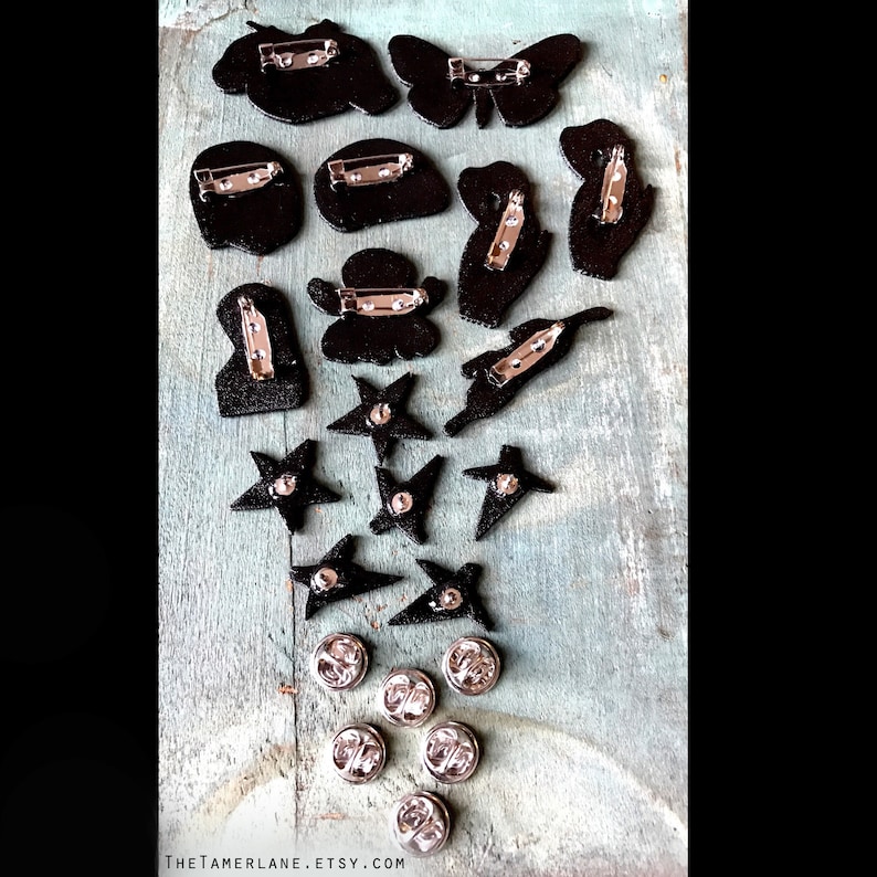 May include: A collection of 15 black and silver pins featuring various designs, including butterflies, stars, and abstract shapes. The pins are displayed on a wooden surface.