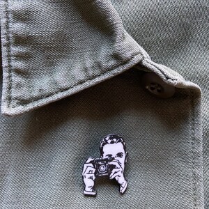 Puede incluir: Pin de esmalte blanco y negro de una persona sosteniendo una c&aacute;mara. El pin est&aacute; sobre un fondo de tela verde.