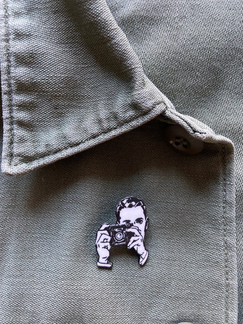 Puede incluir: Pin de esmalte blanco y negro de una persona sosteniendo una c&aacute;mara. El pin est&aacute; sobre un fondo de tela verde.
