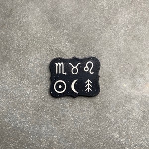 May include: Black and white zodiac sign and celestial symbol design on a black leather coaster. The symbols include the zodiac signs for Scorpio, Taurus, and Leo, as well as a sun, moon, and a pine tree.