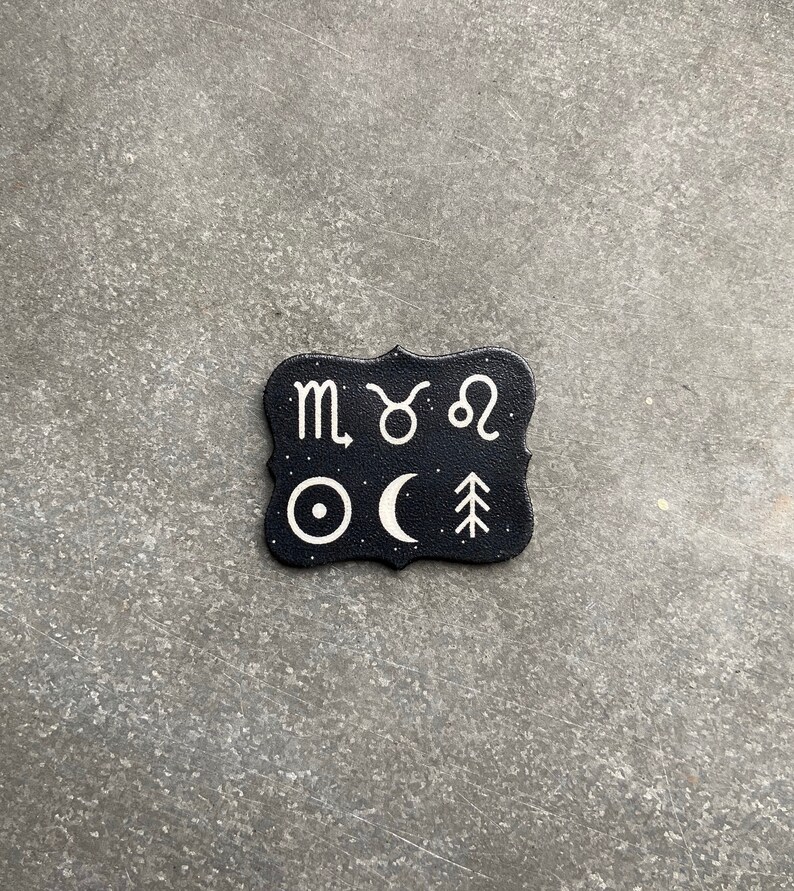 May include: Black and white zodiac sign and celestial symbol design on a black leather coaster. The symbols include the zodiac signs for Scorpio, Taurus, and Leo, as well as a sun, moon, and a pine tree.