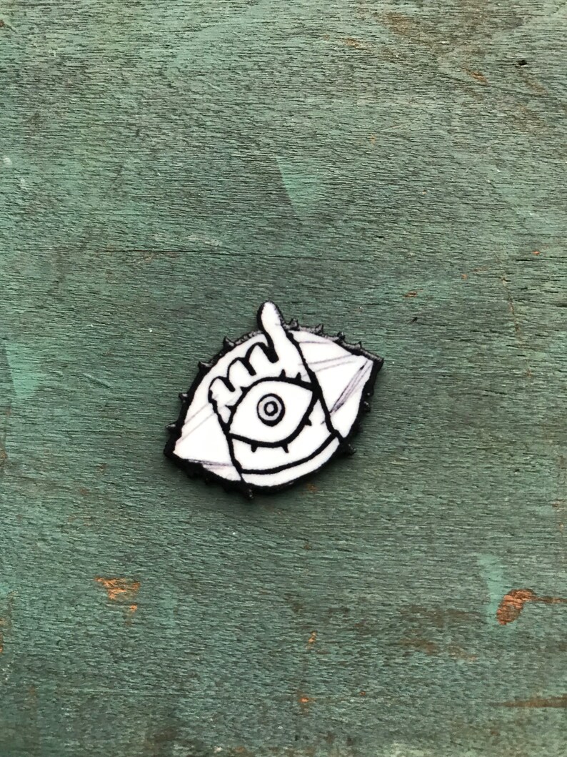 May include: A black and white enamel pin featuring an eye with a hand pointing upwards. The eye is surrounded by a circle of spikes.