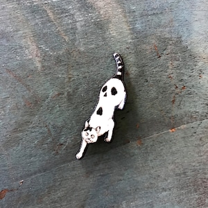 May include: A black and white wooden cat pin with a skull design on its back. The cat is in a crouching position with its tail curled up.