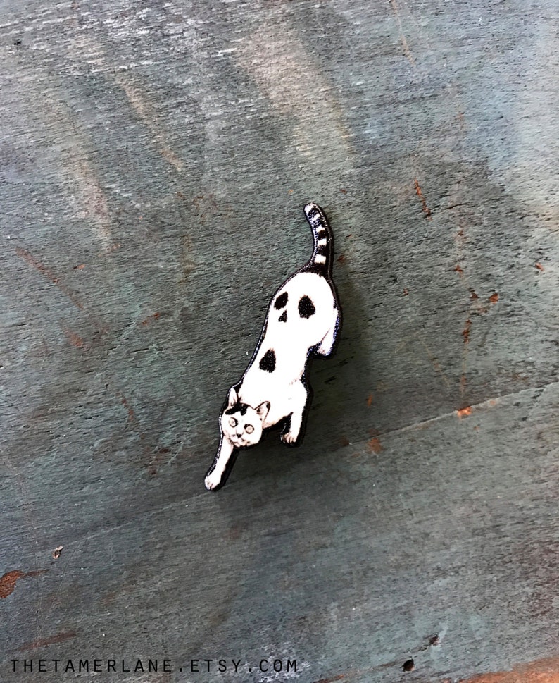 Puede incluir: Un pin de madera de gato blanco y negro con un dise&ntilde;o de calavera en la espalda. El gato est&aacute; en posici&oacute;n de cuclillas con la cola enroscada.