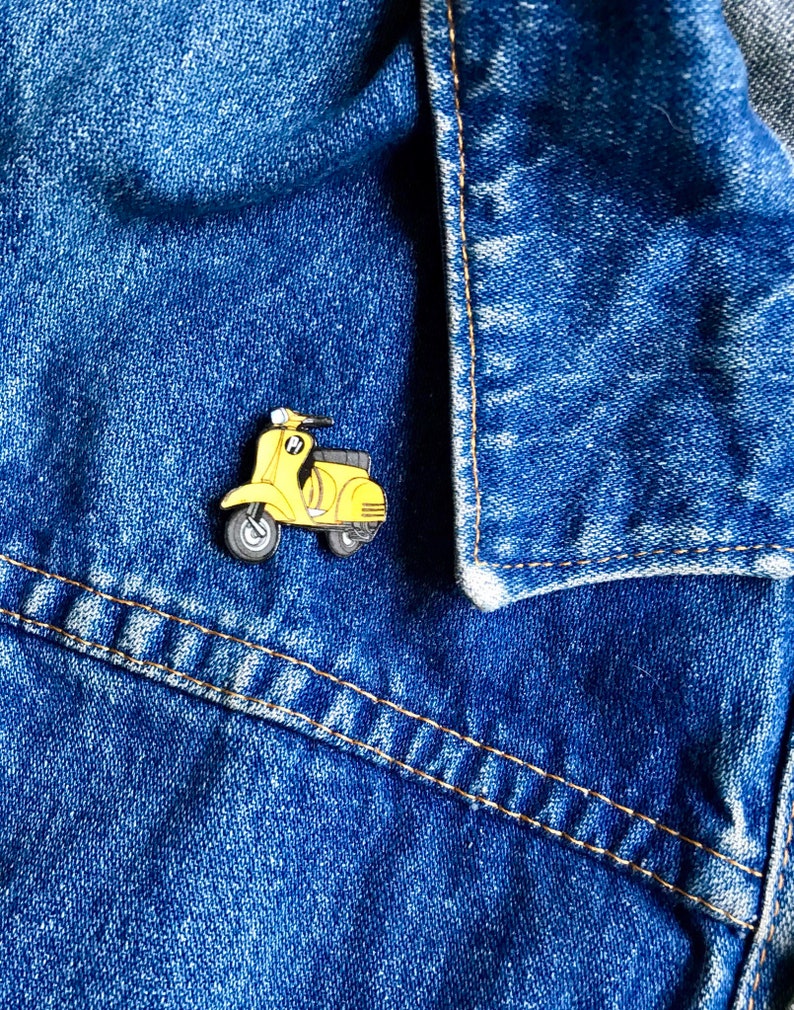 May include: A yellow enamel pin of a vintage scooter. The pin is on a blue denim jacket.