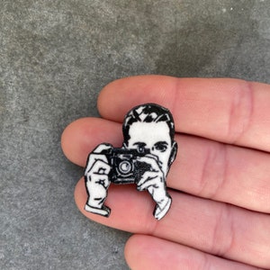 Puede incluir: Pin de esmalte en blanco y negro de una persona sosteniendo una c&aacute;mara. El pin es un dise&ntilde;o en 3D y tiene un acabado brillante.