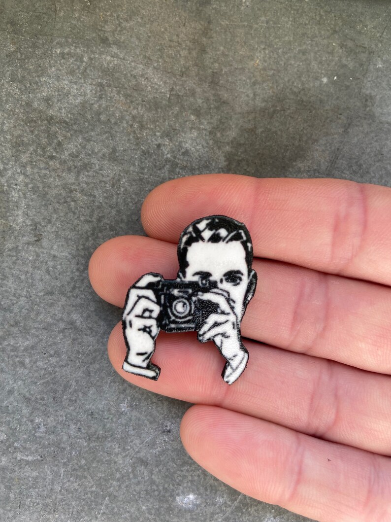 Puede incluir: Pin de esmalte en blanco y negro de una persona sosteniendo una c&aacute;mara. El pin es un dise&ntilde;o en 3D y tiene un acabado brillante.