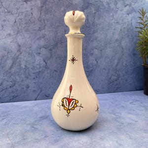 Op de afbeelding: Witte keramische fles met een bloemenmotief in rood, geel en zwart. De fles heeft een decoratieve dop met een wit en gouden ontwerp.