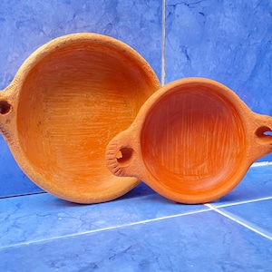 Puede incluir: Dos cuencos de terracota con asas. El cuenco más grande es naranja y el más pequeño es de un tono naranja ligeramente más oscuro.