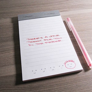 May include: A white notepad with red handwriting that says "There's a little treat for you in the fridge." There are also several grey smiley faces drawn at the bottom of the page.