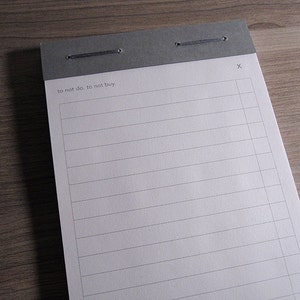 May include: A gray lined notepad with a gray header and a black string tied around the top. The header has the text "to not do. to not buy." and an "X" in the top right corner.