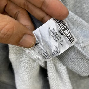 Peut inclure: Gros plan sur une &eacute;tiquette de sweat-shirt grise avec le texte "SESAME STREET" dans un cadre rectangulaire noir. L'&eacute;tiquette comprend &eacute;galement le texte "KAWS" et "All Rights Reserved."