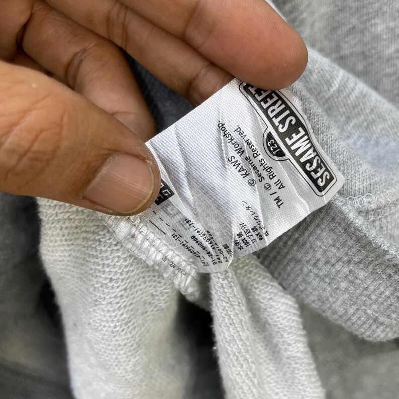 Peut inclure: Gros plan sur une &eacute;tiquette de sweat-shirt grise avec le texte "SESAME STREET" dans un cadre rectangulaire noir. L'&eacute;tiquette comprend &eacute;galement le texte "KAWS" et "All Rights Reserved."
