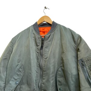 Puede incluir: Una chaqueta bomber verde salvia con cuello gris y forro naranja. La chaqueta tiene cierre de cremallera completo, dos bolsillos delanteros y un bolsillo con cremallera en la manga izquierda. Cuelga de una percha de madera.