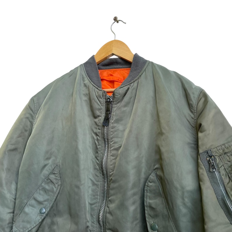 Puede incluir: Una chaqueta bomber verde salvia con cuello gris y forro naranja. La chaqueta tiene cierre de cremallera completo, dos bolsillos delanteros y un bolsillo con cremallera en la manga izquierda. Cuelga de una percha de madera.