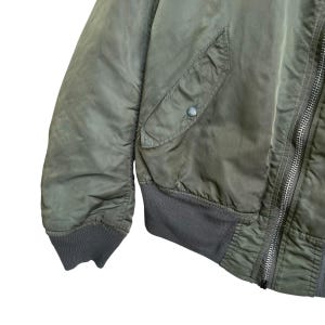 Puede incluir: Primer plano de una chaqueta bomber verde oliva. La chaqueta tiene dobladillo y pu&ntilde;os de canal&eacute;, cierre de cremallera y un bolsillo con bot&oacute;n. La tela tiene un ligero brillo.