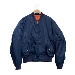 May include: A navy blue bomber jacket with a zip-up front and two front pockets. The jacket has a ribbed collar, cuffs, and hem. The interior lining is orange. The jacket is hanging from a wooden hanger.