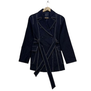 May include: A dark blue denim jacket with white stitching and a double-breasted design. The jacket has a matching belt and two front pockets. The jacket is hanging on a wooden hanger.