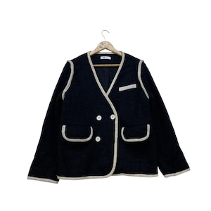 May include: Black jacket with cream-colored trim and two front pockets. The jacket has a double-breasted design with two rows of white buttons. The jacket is hanging on a wooden hanger.
