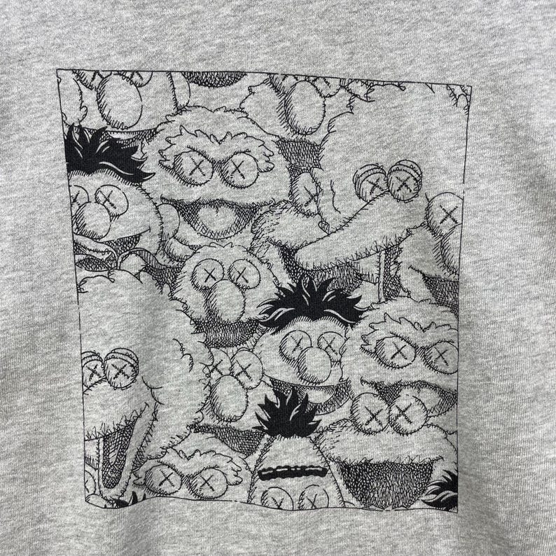 Peut inclure: Un sweat-shirt gris avec un motif noir et blanc de personnages de dessins anim&eacute;s aux yeux en forme de X. Le motif est contenu dans un rectangle noir. Les personnages ont des expressions vari&eacute;es et sont dens&eacute;ment regroup&eacute;s.