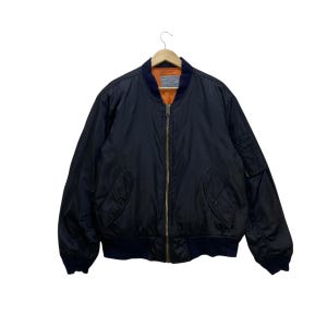 Peut inclure: Veste de bombardier noire avec col et poignets bleu marine, suspendue à un cintre en bois. La veste a une fermeture éclair et deux poches avant. La doublure intérieure est orange.