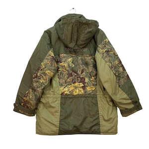 Peut inclure: Veste à capuche vert olive avec un motif floral au dos et sur les manches. La veste a une capuche à cordon de serrage, des poignets boutonnés et le mot "courmayeur" brodé dans le haut du dos.