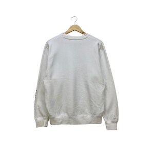 Puede incluir: Una sudadera blanca de manga larga colgada de una percha de madera. La sudadera tiene cuello redondo y pu&ntilde;os y dobladillo de canal&eacute;. Hay texto en la manga izquierda. El fondo es blanco.