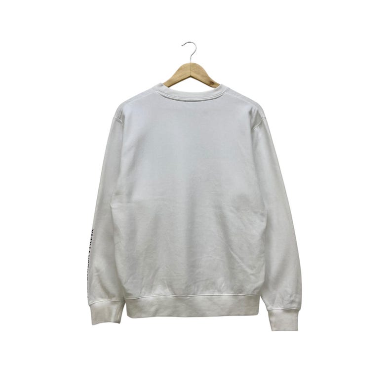 Puede incluir: Una sudadera blanca de manga larga colgada de una percha de madera. La sudadera tiene cuello redondo y pu&ntilde;os y dobladillo de canal&eacute;. Hay texto en la manga izquierda. El fondo es blanco.