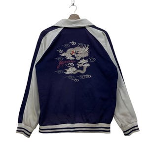 Puede incluir: Una chaqueta de béisbol azul marino y blanca con un bordado de dragón y nubes en la espalda. El bordado incluye la palabra "Japón".