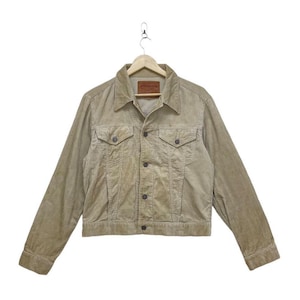 May include: A tan corduroy jacket with a button-down front, two chest pockets, and a collar. The jacket has a small brown label on the chest. The jacket is hanging on a hanger against a white background.