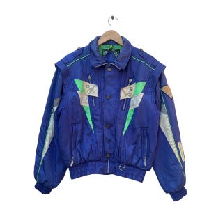 Peut inclure: Veste bleue vintage avec des accents verts et argentés. La veste présente une fermeture éclair, des boutons et un ourlet côtelé. Le design comprend des détails verts et argentés en forme d'éclair sur le devant et les manches. La veste est suspendue à un cintre en bois.