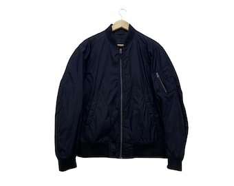 Puede incluir: Chaqueta bomber azul marino con cremallera, puños y dobladillo acanalados. La chaqueta tiene un pequeño bolsillo en la manga izquierda y está colgada de una percha de madera. La chaqueta está hecha de un material ligero.