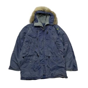 May include: A navy blue winter parka with a faux fur-lined hood. The jacket has a button and zip fastening, multiple pockets, and a small embroidered emblem on the sleeve. The parka is designed for cold weather.