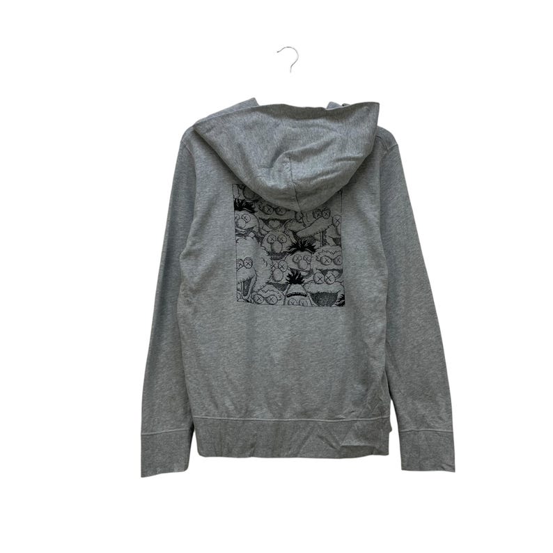 Peut inclure: Un sweat &agrave; capuche gris zipp&eacute; avec une capuche. Le dos du sweat &agrave; capuche pr&eacute;sente une impression graphique en noir et blanc de personnages de dessins anim&eacute;s. Le sweat &agrave; capuche est suspendu &agrave; un crochet blanc sur fond blanc.