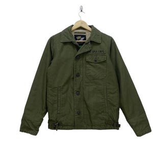 May include: Olive green jacket with a button-up front, two chest pockets, and a collared neckline. The jacket has the text "FAT SPLY CAPS" on the right chest. The jacket is hanging on a wooden hanger.