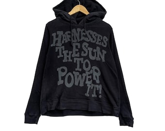 May include: Black hoodie with a drawstring hood and long sleeves. The front of the hoodie features the text "HARNESSES THE SUN TO POWER IT!" in a gray, distressed font. The hoodie is hanging on a wooden hanger.