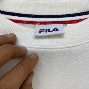 Puede incluir: Primer plano de un cuello de sudadera blanco con un ribete a rayas azules, rojas y blancas. El logotipo de Fila es visible en una etiqueta rectangular. La tela tiene una textura acanalada.