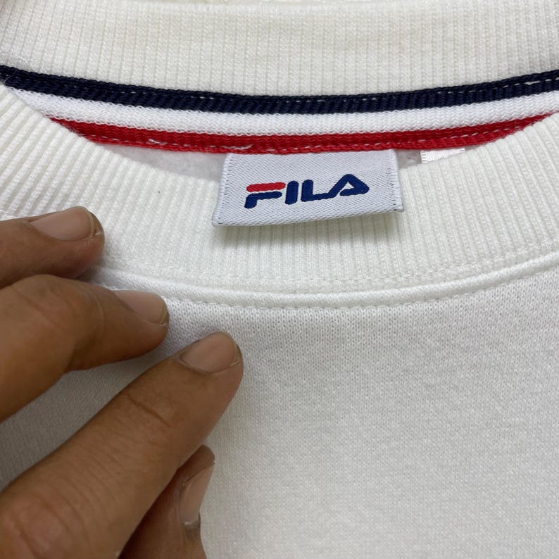 Puede incluir: Primer plano de un cuello de sudadera blanco con un ribete a rayas azules, rojas y blancas. El logotipo de Fila es visible en una etiqueta rectangular. La tela tiene una textura acanalada.