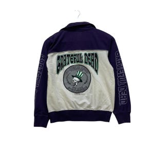 May include: A navy blue and white zip-up jacket featuring the words "Grateful Dead" in green and a skull wearing a top hat. The sleeves have the words "Grateful Dead" printed on them. The jacket is hanging from a white hanger.