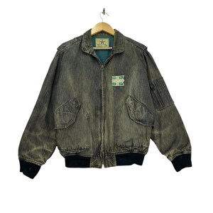 May include: A charcoal grey acid-wash denim bomber jacket with a full-length front zipper, ribbed black cuffs and hem, two large front flap pockets, and a utility pocket on the left sleeve. A green patch on the chest features the text STAFF.