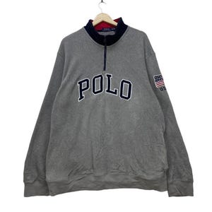 May include: A gray fleece pullover sweatshirt with a navy blue zipper and a navy blue "POLO" logo on the front. The sweatshirt has a small American flag patch on the right sleeve.