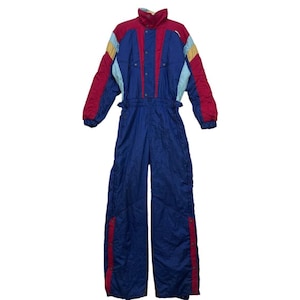 Peut inclure: Combinaison de ski vintage bleue et rouge avec des accents de blocs de couleurs. La combinaison une pièce présente un col montant, des fermetures à pression et des rayures rouges sur les jambes. Les manches ont des empiècements jaunes et bleu clair.