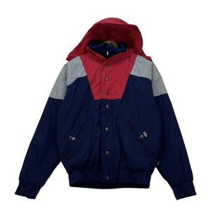 Peut inclure: Veste à capuche avec un design color block. La veste présente une capuche et le haut de la poitrine rouges, une section d'épaule grise et un corps bleu marine. Elle a une fermeture à boutons et deux poches.