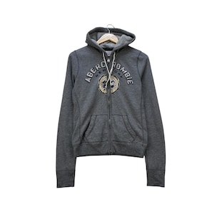 May include: A grey zip-up hoodie featuring the Abercrombie New York logo on the chest. The hoodie has a drawstring hood, front pockets, and ribbed cuffs. The logo is embroidered in a circular design with gold and navy blue accents.