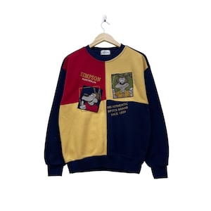 Puede incluir: Una sudadera vintage de Simpson Australia. El diseño de bloques de color presenta paneles rojos, amarillos y azul marino. La sudadera tiene gráficos de dibujos animados y el texto "SIMPSON AUSTRALIA" y "THE AUTHENTIC SPORTS BRAND SINCE 1939".